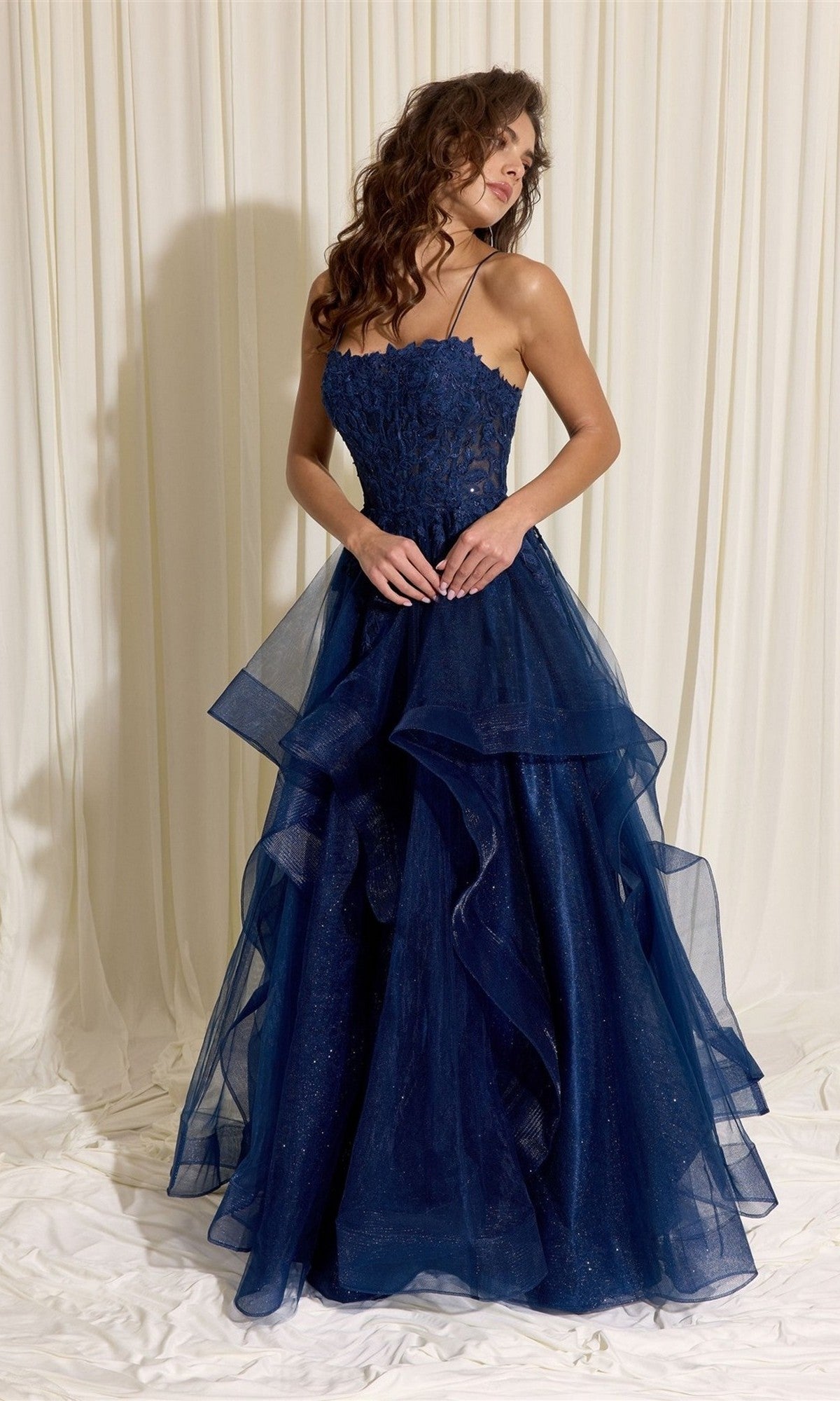 A woman wears the Eureka EK167 Long Prom Dress, featuring a navy blue floral lace corset bodice and a layered tulle A-line skirt, standing against a cream-colored fabric backdrop.